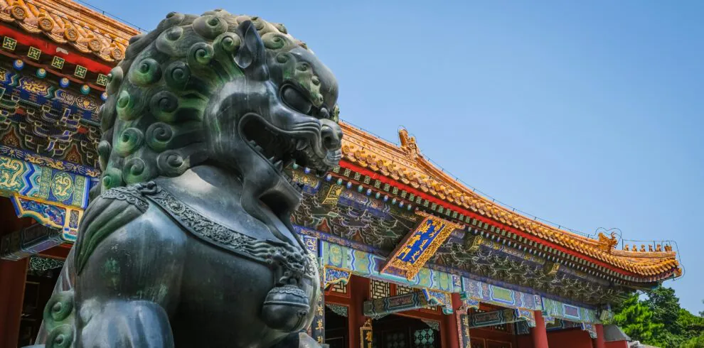 Forbidden City, Beijing, Chinatravel, Bronze guardian lion statue in front of a traditional Chinese temple with ornate, colorful roof tiles against a clear blue sky.
