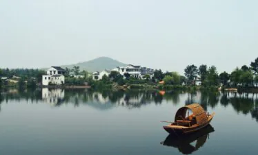 Chinatrip, Calm lakeside scene with white houses along the shore, green trees, and a wooden boat in the foreground on a reflective lake.