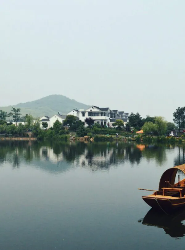 Chinatrip, Calm lakeside scene with white houses along the shore, green trees, and a wooden boat in the foreground on a reflective lake.