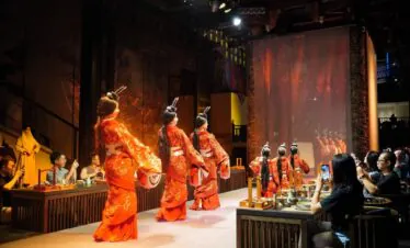 sichuan, china, china tour, Group of dancers in orange traditional outfits performing on stage while audience films with smartphones nearby strain their attention to the performance.