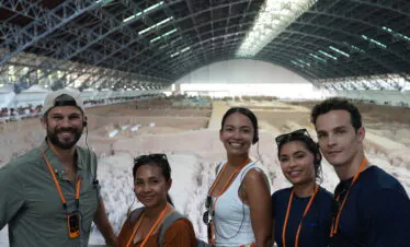 Terra Cotta Warriors, xi'an, china, china tour, Five people smiling for a selfie at an indoor archaeological dig, with a large curved roof and excavated area behind them, all wearing orange lanyards and headsets.