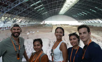 Terra Cotta Warriors, xi'an, china, china tour, Five people smiling for a selfie at an indoor archaeological dig, with a large curved roof and excavated area behind them, all wearing orange lanyards and headsets.