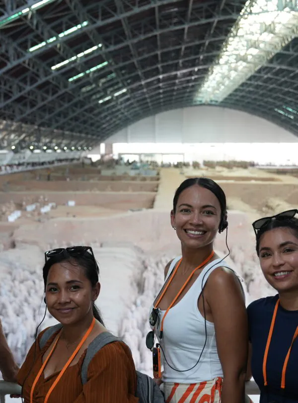 Terra Cotta Warriors, xi'an, china, china tour, Five people smiling for a selfie at an indoor archaeological dig, with a large curved roof and excavated area behind them, all wearing orange lanyards and headsets.