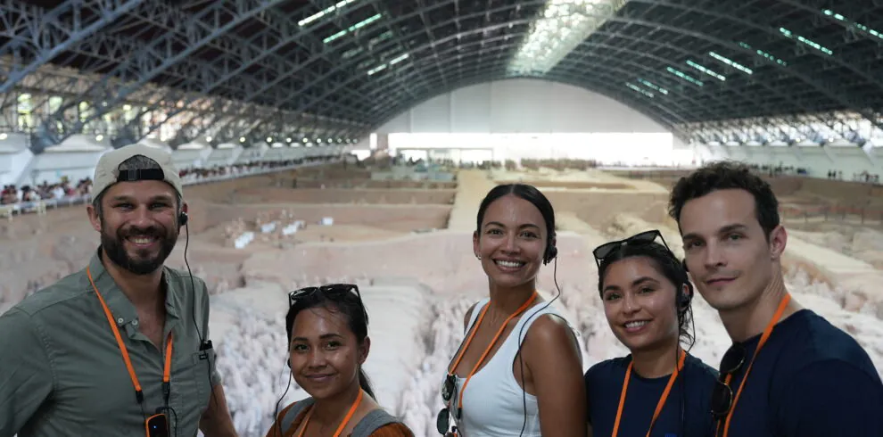 Terra Cotta Warriors, xi'an, china, china tour, Five people smiling for a selfie at an indoor archaeological dig, with a large curved roof and excavated area behind them, all wearing orange lanyards and headsets.