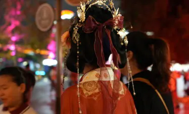hanfu, xi'an, china, china tour, Back view of a woman in a red traditional outfit wearing an ornate gold headpiece with a red ribbon at a nighttime festival outside the city.