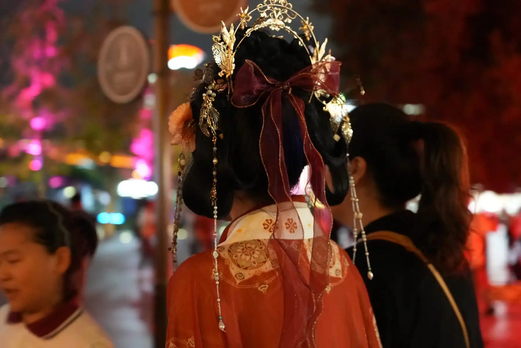 hanfu, xi'an, china, china tour, Back view of a woman in a red traditional outfit wearing an ornate gold headpiece with a red ribbon at a nighttime festival outside the city.