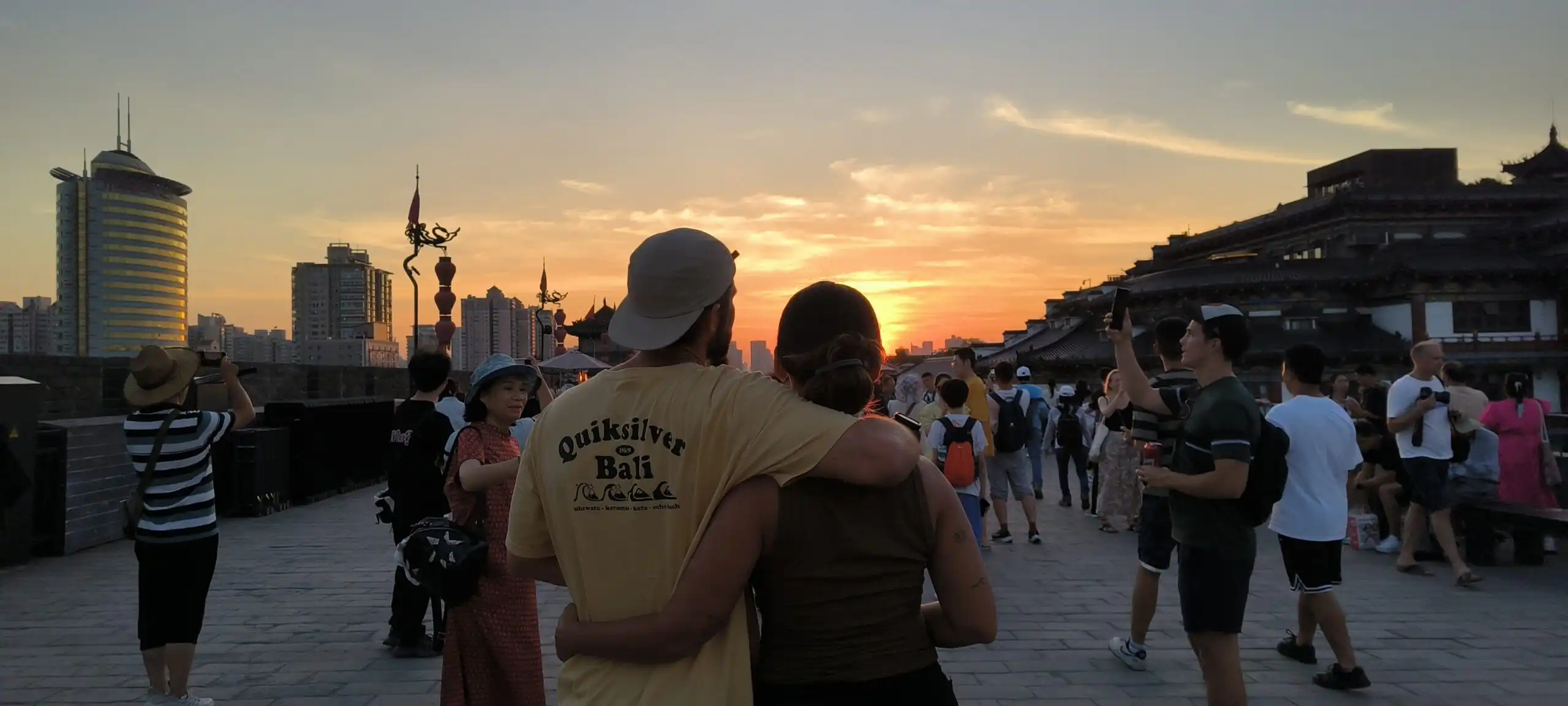 xi'an, china, china tour, Couple embraces on a historic city wall at sunset, with a crowd and modern high-rises in the background.Months tone can vary, but keep to the sense of scene and mood.
