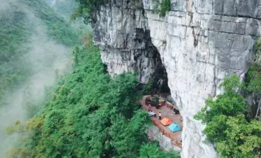 Cave Camping in Guizhou, china, china tour, Cliffside campsite on a rock face with orange and blue tents and seating on a wooden platform amid a misty forest valley.