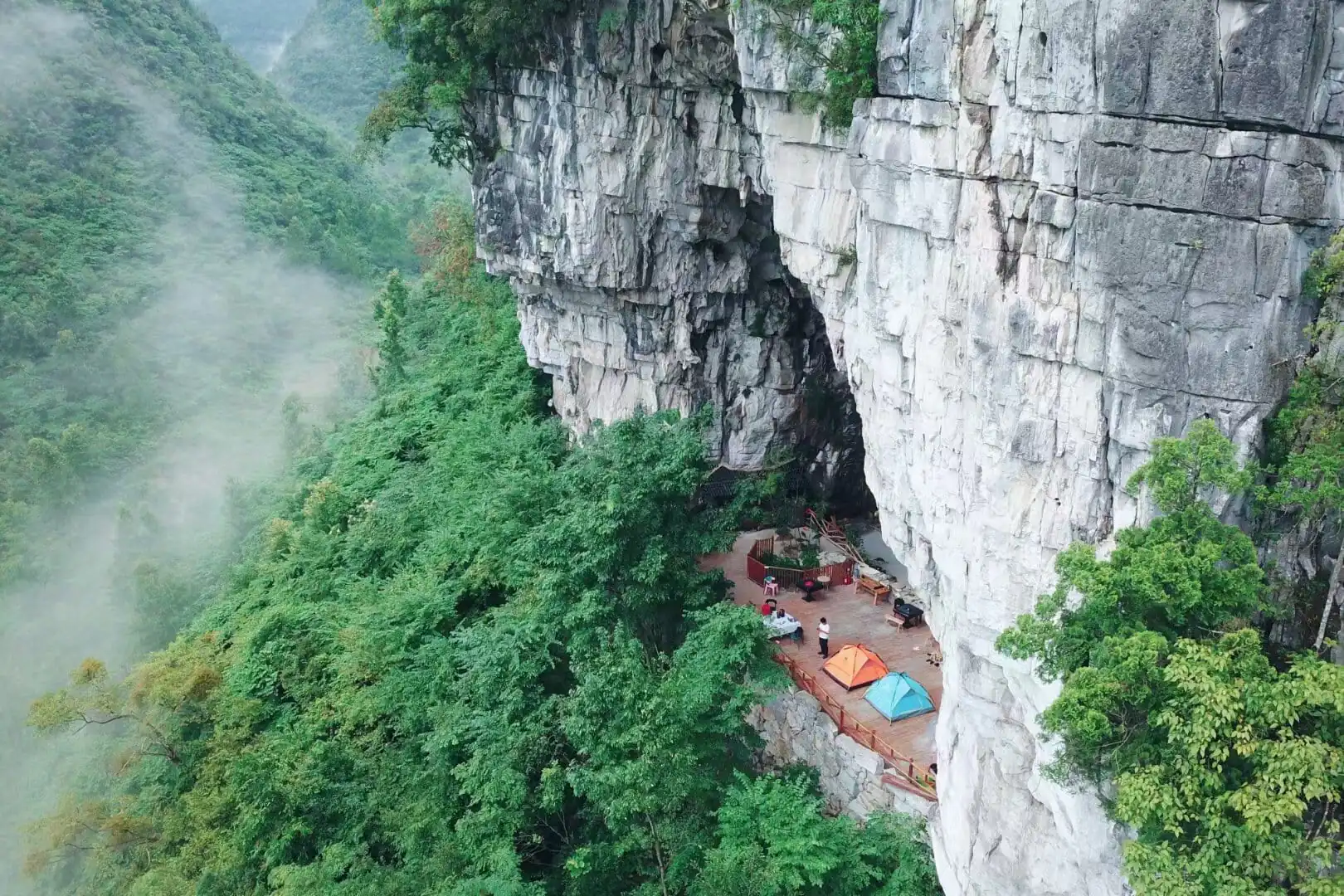 Cave Camping in Guizhou, china, china tour, Cliffside campsite on a rock face with orange and blue tents and seating on a wooden platform amid a misty forest valley.