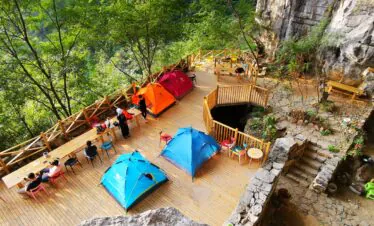 Cave Camping in Guizhou, china, china tour, Cliffside wooden deck cafe with colorful tents, tables, and chairs surrounded by trees and rocky ledges.