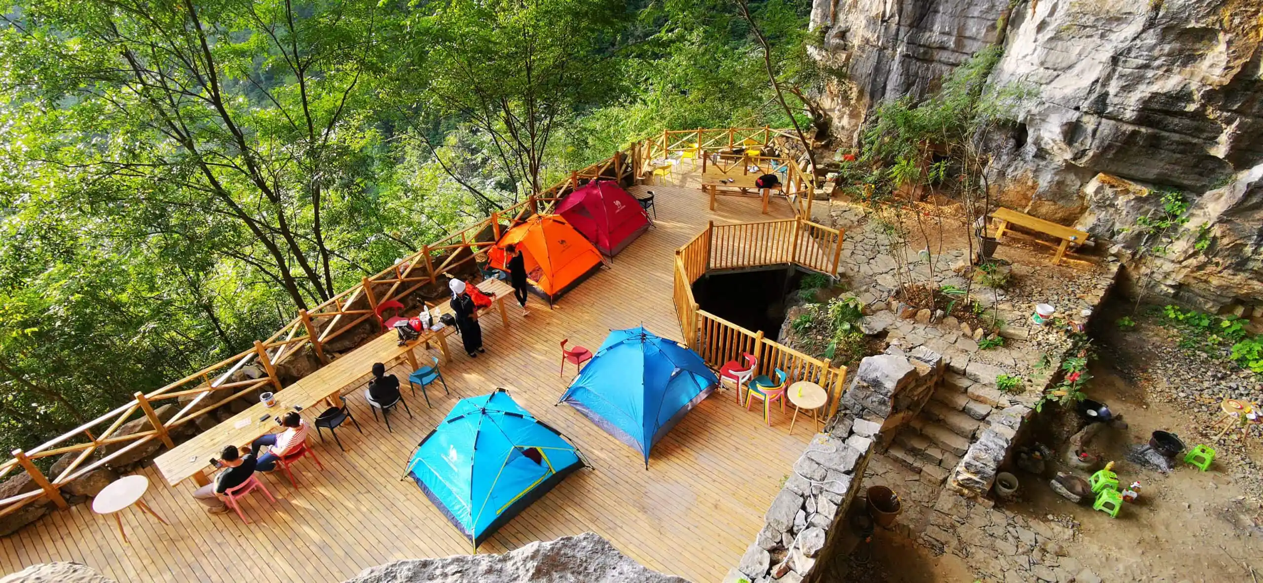 Cave Camping in Guizhou, china, china tour, Cliffside wooden deck cafe with colorful tents, tables, and chairs surrounded by trees and rocky ledges.