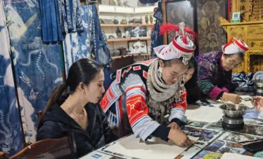 Batik experience in Miao minority, Guizhou, china, china tour, People in traditional dress at a craft workshop, writing or painting at a table with mirrors and printed sheets in a shop filled with blue fabrics and curiosities.