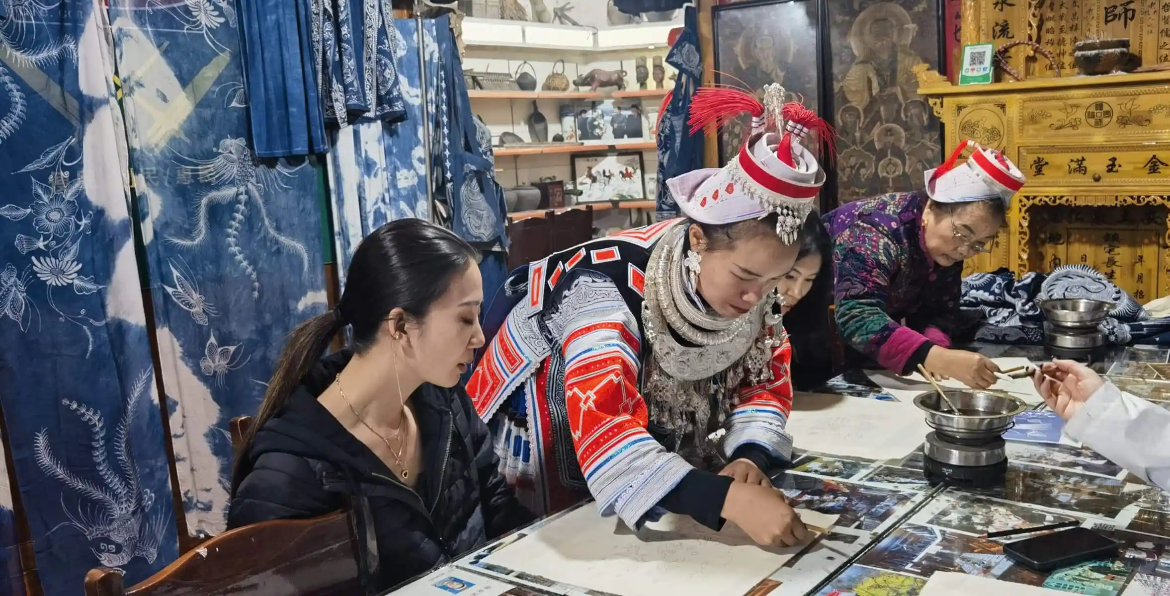 Batik experience in Miao minority, Guizhou, china, china tour, People in traditional dress at a craft workshop, writing or painting at a table with mirrors and printed sheets in a shop filled with blue fabrics and curiosities.