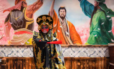 A performer in elaborate black and gold Chinese opera robes with a red painted face stands with a red prop in front of a mural of warriors and lotus blossoms.