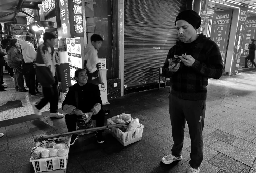 dining culture, Chinese Festivals: How to Appreciate Food Culture, chinaexplorertour, Black-and-white street scene: a man in a beanie eats from a bowl on the sidewalk while a street vendor sits nearby with baskets of bread and goods; illuminated signs and pedestrians in the background.