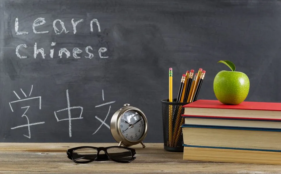 How to Overcome Language Barriers in China: Basic Chinese, chinaexplorertour, Student desk scene for language learning: chalkboard reads 'Learn Chinese' with Chinese characters, pencils, clock, glasses, and a stack of books with a green apple on top