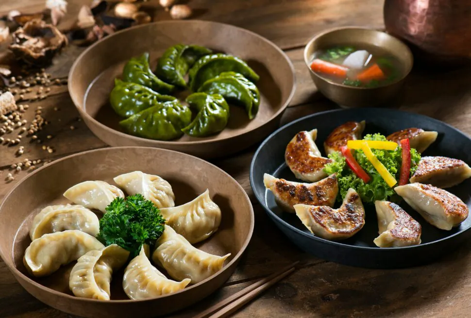 Dumplings, Chinese food, chinaexplorertour, Assorted dumplings on a wooden table: green spinach dumplings in a brown bowl, crescent-shaped boiled dumplings with parsley, and pan-fried dumplings with vegetables on a dark plate, with a small soup bowl in the background.