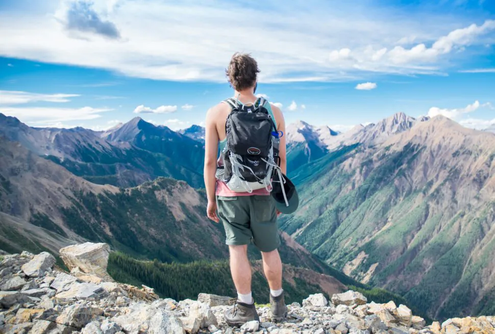 How to Travel in China on a Budget: Money-Saving Tips, chinaexplorertour, Back view of a hiker standing on a rocky ridge, wearing a backpack and shorts, looking at snowless mountain peaks under a blue sky.
