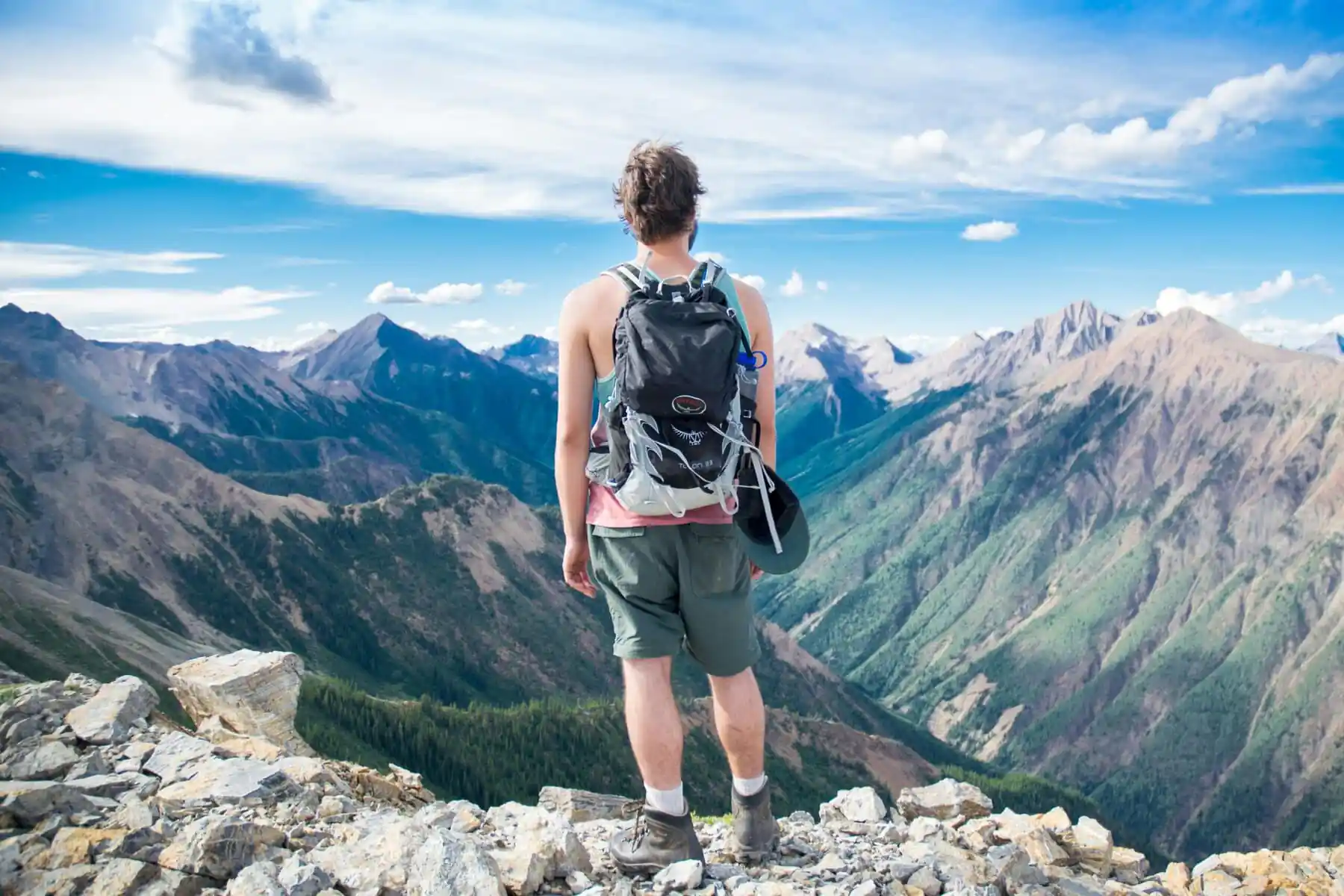 How to Travel in China on a Budget: Money-Saving Tips, chinaexplorertour, Back view of a hiker standing on a rocky ridge, wearing a backpack and shorts, looking at snowless mountain peaks under a blue sky.