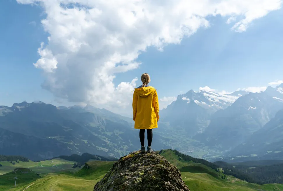 How to Travel Solo in Your China Tour, chinaexplorertour, Person in a yellow jacket stands on a rocky peak, gazing at a valley and snow-capped mountains under a blue sky.