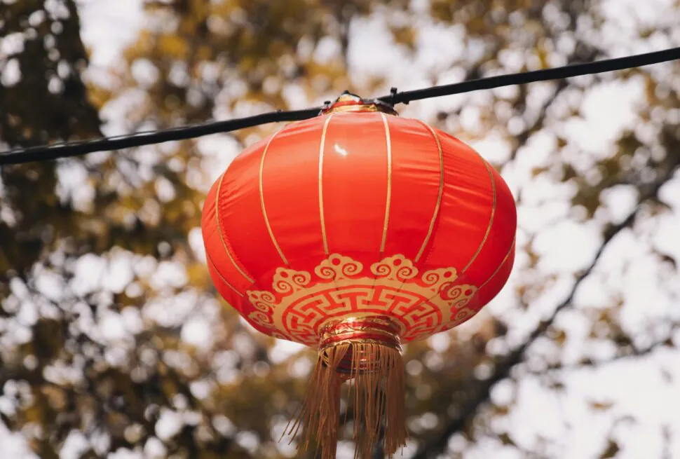 Red Chinese lantern hanging outdoors with gold patterns and tassels, blurred autumn foliage in the background.
