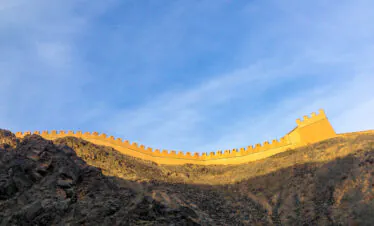 Curved stone fortress wall with crenellations along a rocky hillside under a clear blue sky