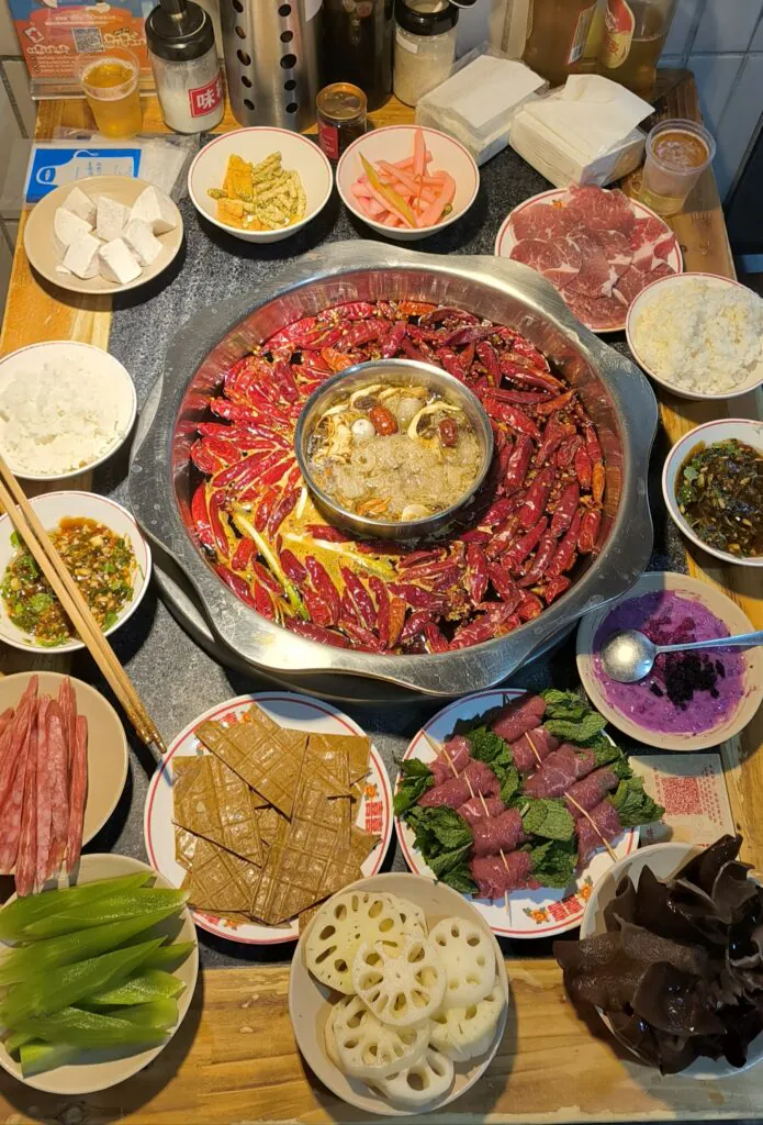 A table set for a traditional Chinese hot pot meal featuring a central pot of spicy broth with red chili peppers. Surrounding it are plates of sliced meats, vegetables, tofu, rice, and dipping sauces.