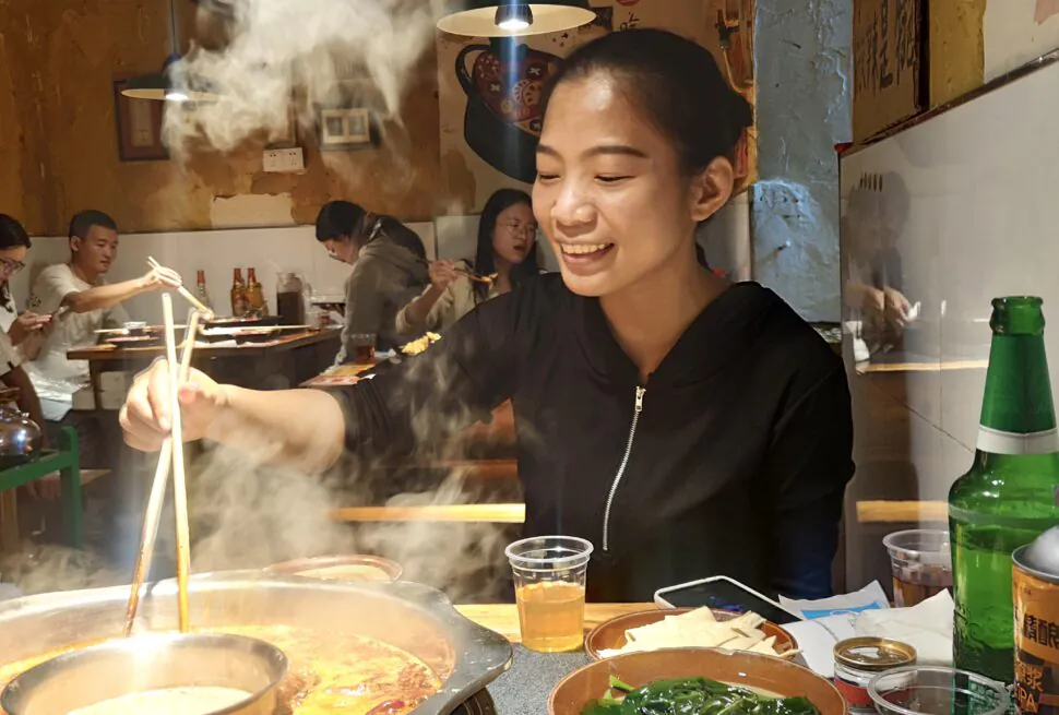 Hotpot experience, sichuan, china, chinaexplorertour, Smiling woman uses chopsticks to cook at a steaming hot pot in a casual restaurant with diners in the background