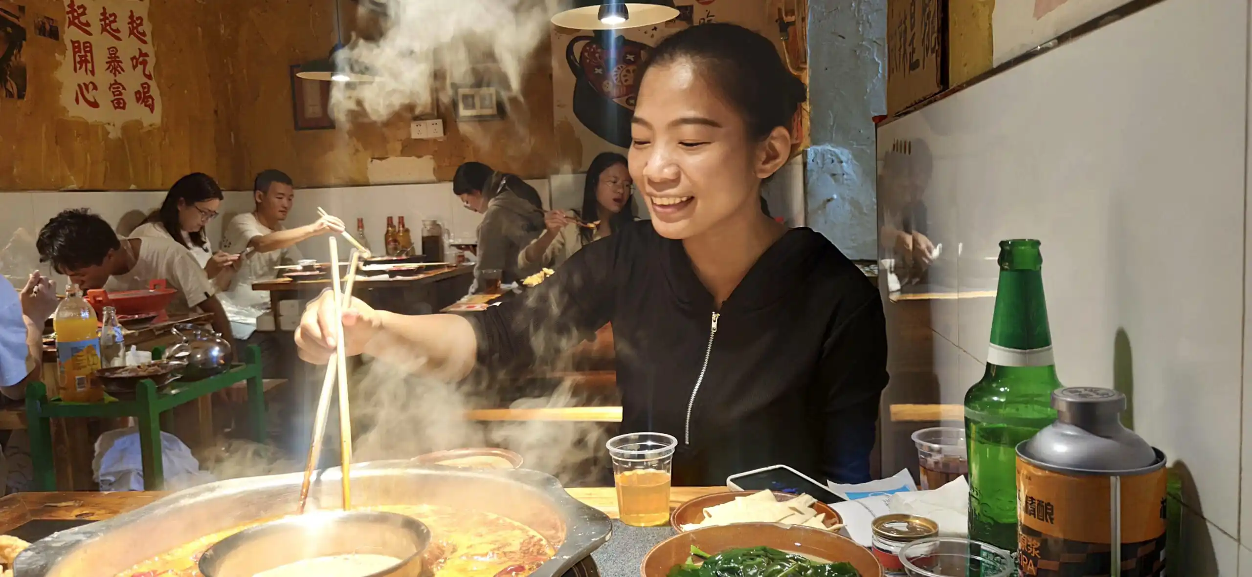 Hotpot experience, sichuan, china, chinaexplorertour, Smiling woman uses chopsticks to cook at a steaming hot pot in a casual restaurant with diners in the background