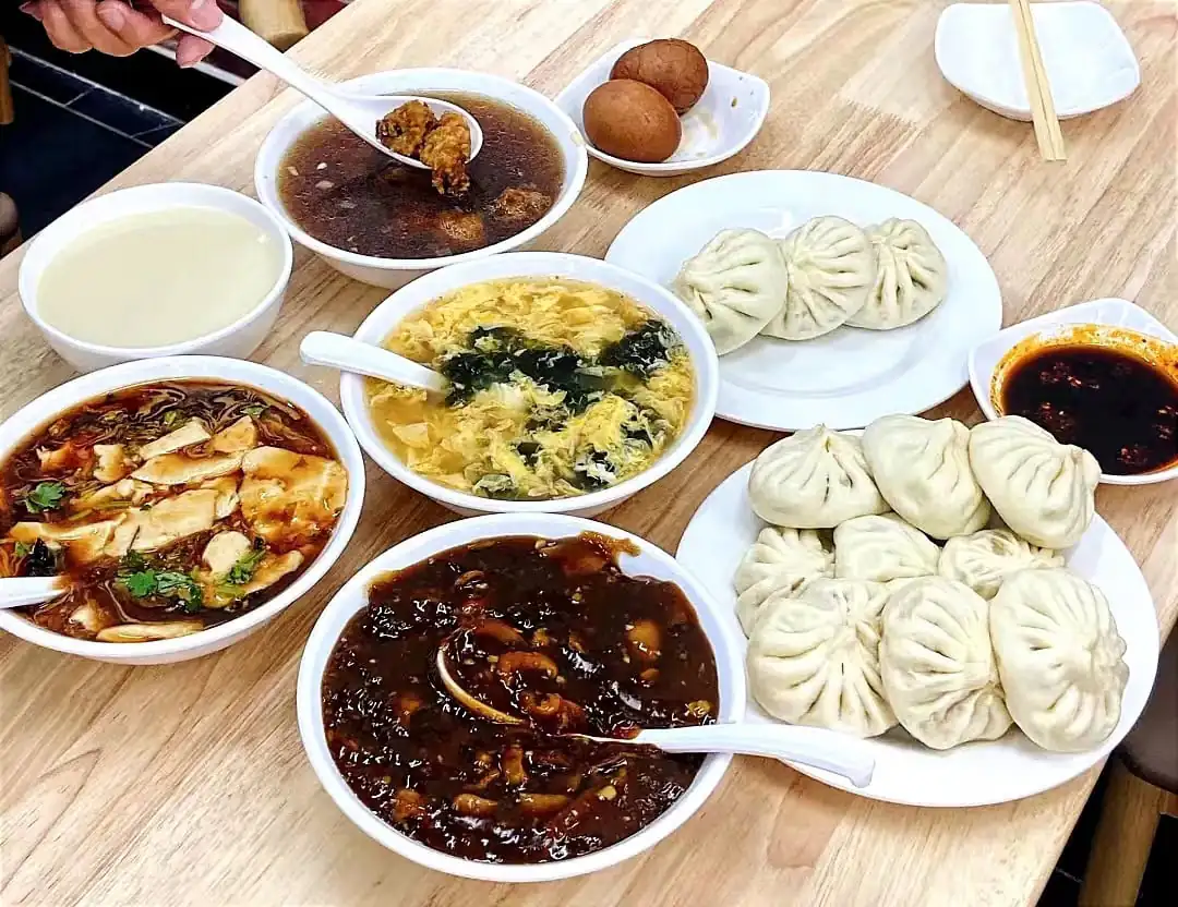 Traditional Chinese Breakfast, china, chinaexplorertour, Steamed dumplings on a white plate with several bowls of soup and sauces on a wooden table.