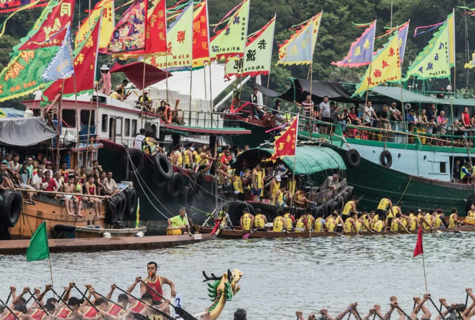 Dragon boat race, china, china tour, Dragon boat race on a river with teams paddling in red and yellow; colorful flags line the boats and a cityscape in the background.