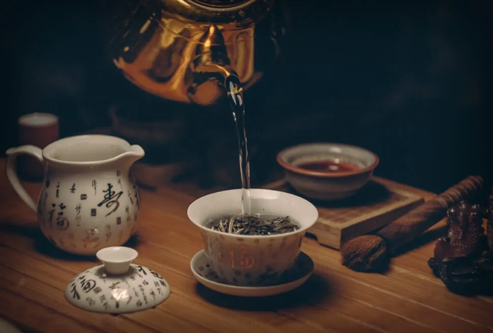 chinese tea, china, china tour, Porcelain teapot with Chinese characters pouring hot water into a teacup on a wooden table, tea leaves visible in the cup.
