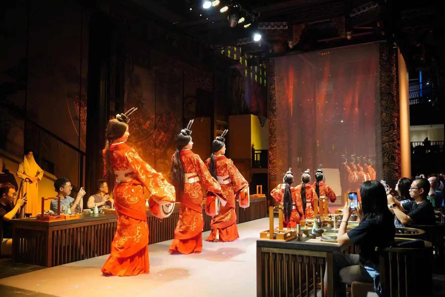 sichuan, china, china tour, Dancers in bright orange, ornate costumes perform a traditional dance on a stage while audience at tables records with phones.