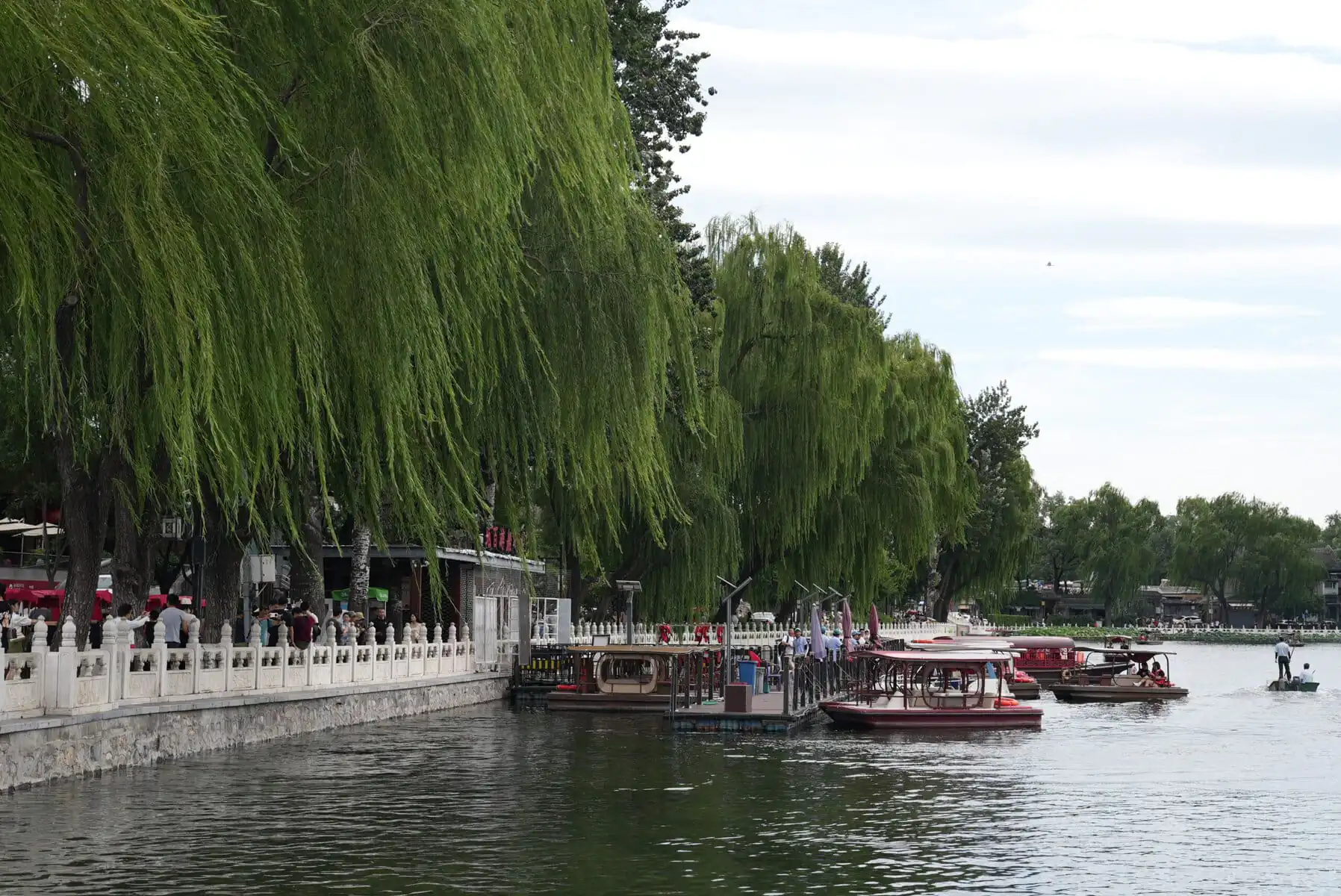 Shichahai, beijing, china, china tour, Riverside promenade with weeping willow trees, a white railing, and people walking by the water with boats docked nearby