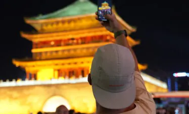 bell tower, xi'an, china, china tour, Person in a light shirt and cap raises a smartphone to photograph a lit golden pagoda at night.