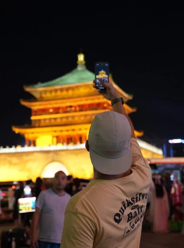 bell tower, xi'an, china, china tour, Person in a light shirt and cap raises a smartphone to photograph a lit golden pagoda at night.