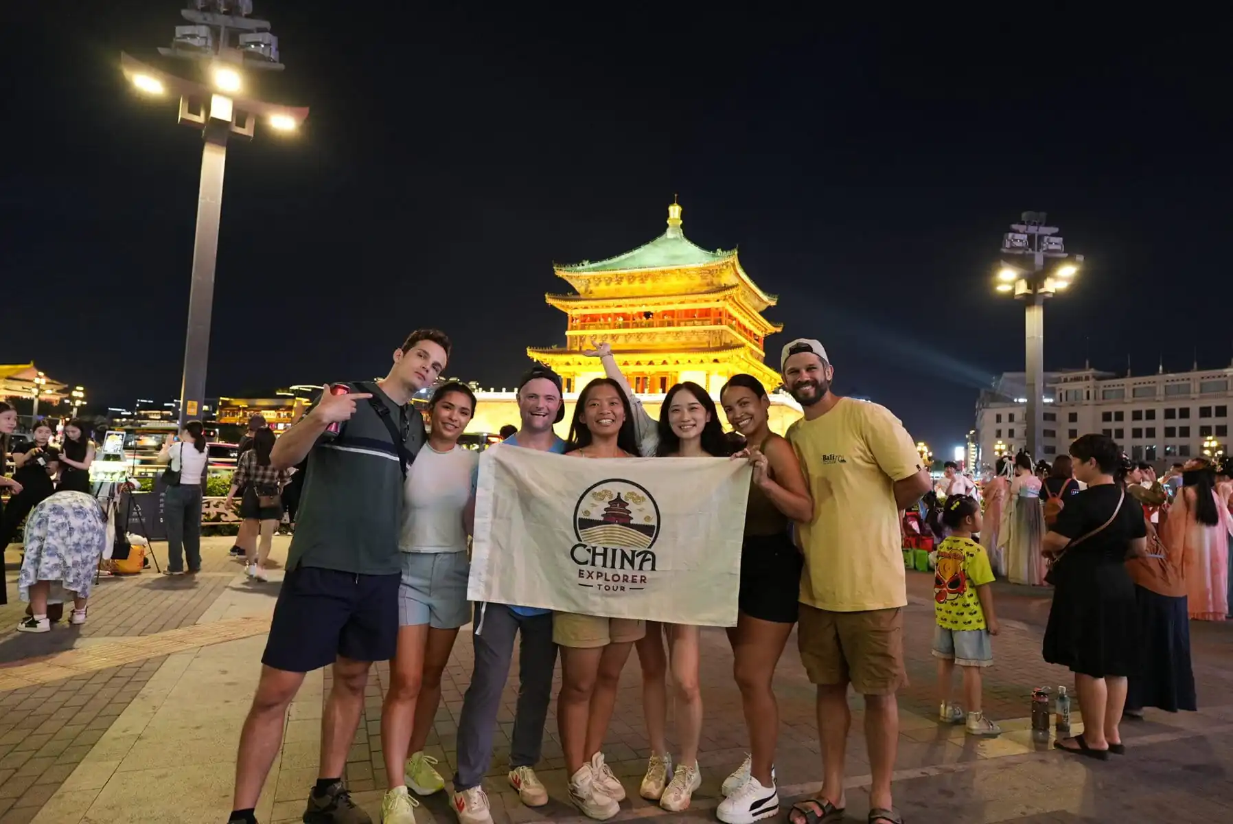 Xi'an Bell Tower with China Explorer Tour, china, china tour, Group of friends posing with a banner that reads 'China Explorer Tour' in front of a lit traditional pagoda at night.
