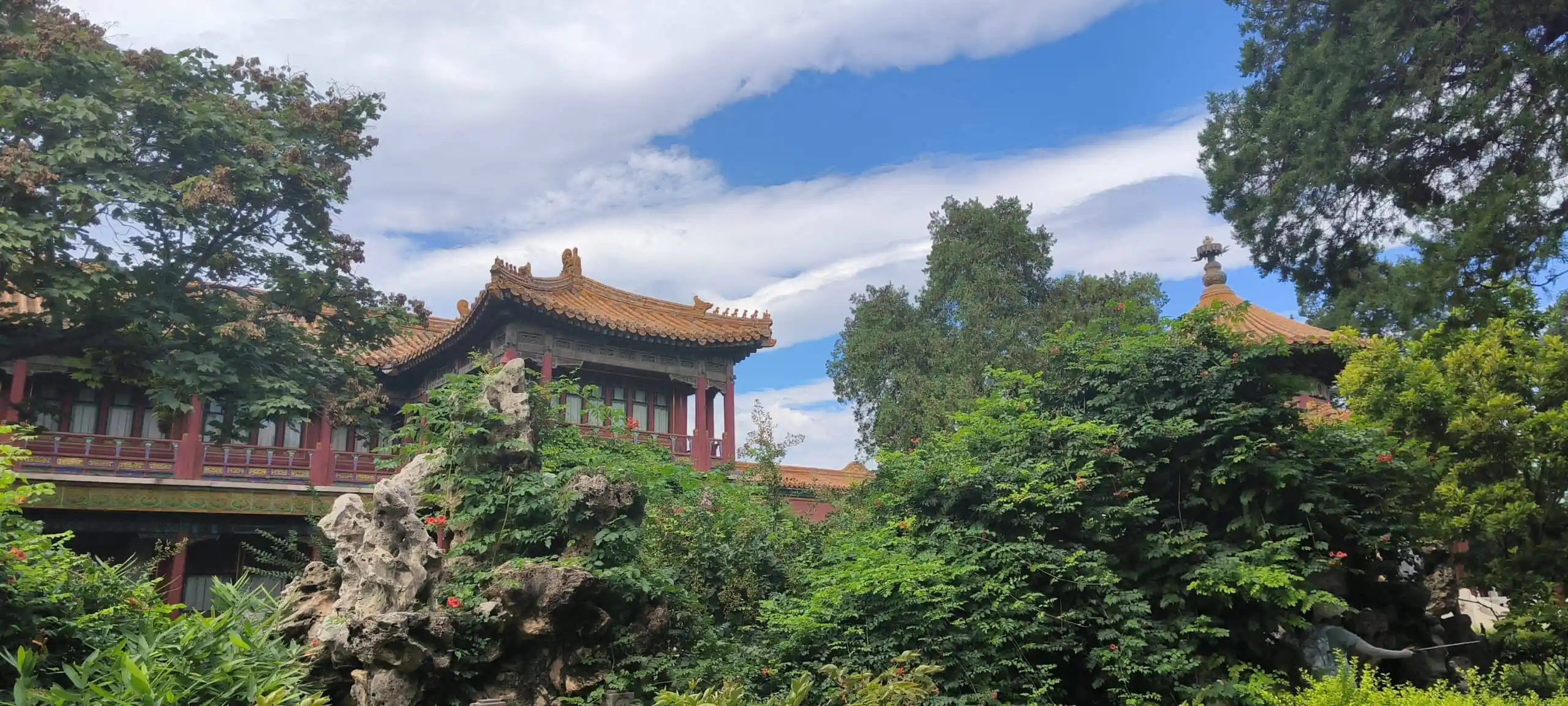 the forbidden city, beijing, china, china tour, Traditional Chinese temple with orange tiled roofs framed by lush greenery and a blue sky.Reflective garden rocks and red wooden beams visible in the foreground, surrounding the temple.