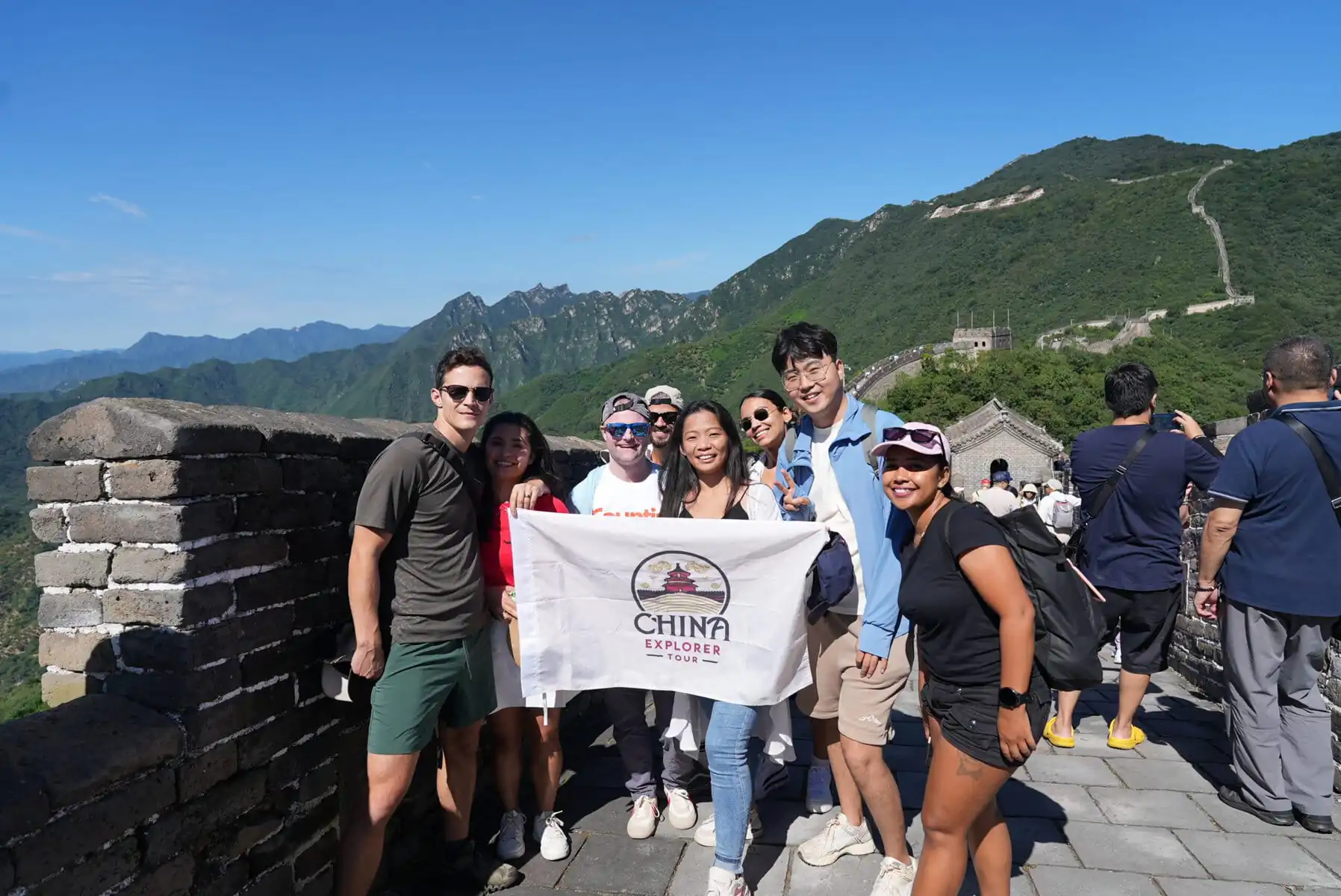 Mutianyu Great Wall, Beijing, Chinatravel, Group of diverse tourists posing with a China Explorer Tour banner on the Great Wall, blue sky and green mountains in the background.