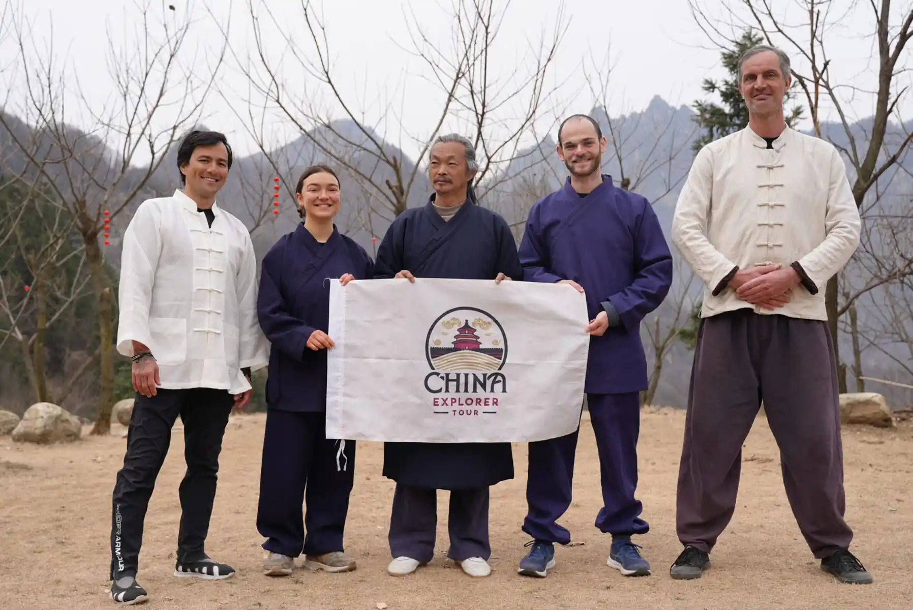 wudang taichi, hubei, china, chinaexplorertour, Five people in martial arts outfits stand outdoors holding a banner that says 'China Explorer Tour' with a mountainous, leafless-tree backdrop.