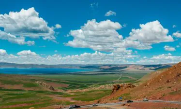 Tibet, china, china tour, Wide valley with green fields, a blue lake in the distance, red rocky hills on the right, and a winding road in the foreground under a bright blue sky with clouds.