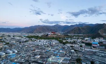 Tibet, china, china tour, City sprawling at the base of brown mountains, with a white/red hillside complex prominent on a hill and a dense grid of low-rise buildings in the foreground. The image shows a large city set against rugged mountains under a blue sky with scattered clouds, and a notable hillside structure in the center-right.