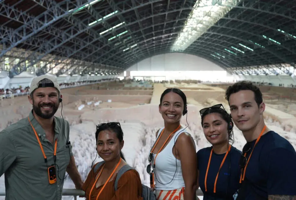 the terracotta army, xi'an, china tour, Five smiling tour guides with headsets and orange lanyards pose together at a large indoor excavation site under a curved steel roof.