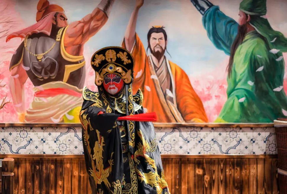 Chinese opera, china tour, Person in an ornate black and gold Chinese opera costume with red mask and headdress, posing with a red prop in front of a mural of warriors.