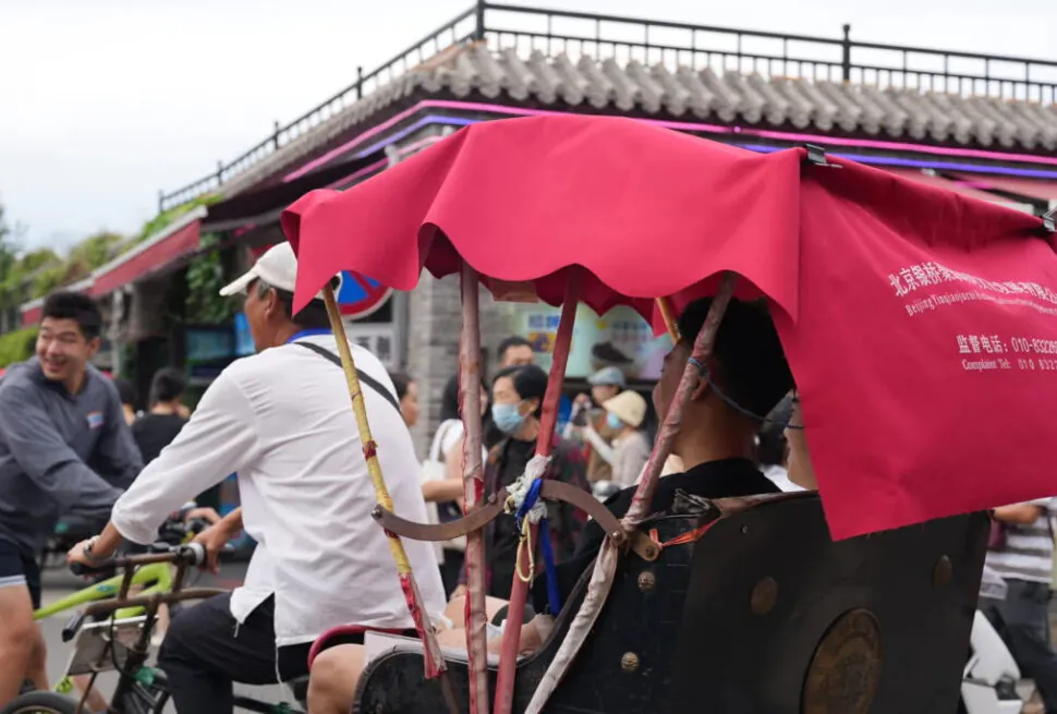 The Importance of Hutongs: Exploring Old Beijing Neighborhoods, china tour, Pedicab with a bright pink canopy carrying a rider, on a busy street with pedestrians and cyclists nearby.