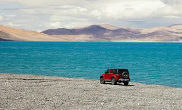 Tibet, china, china tour, Red Jeep parked on a rocky shore beside a bright blue lake with arid mountains in the distance under a cloudy sky