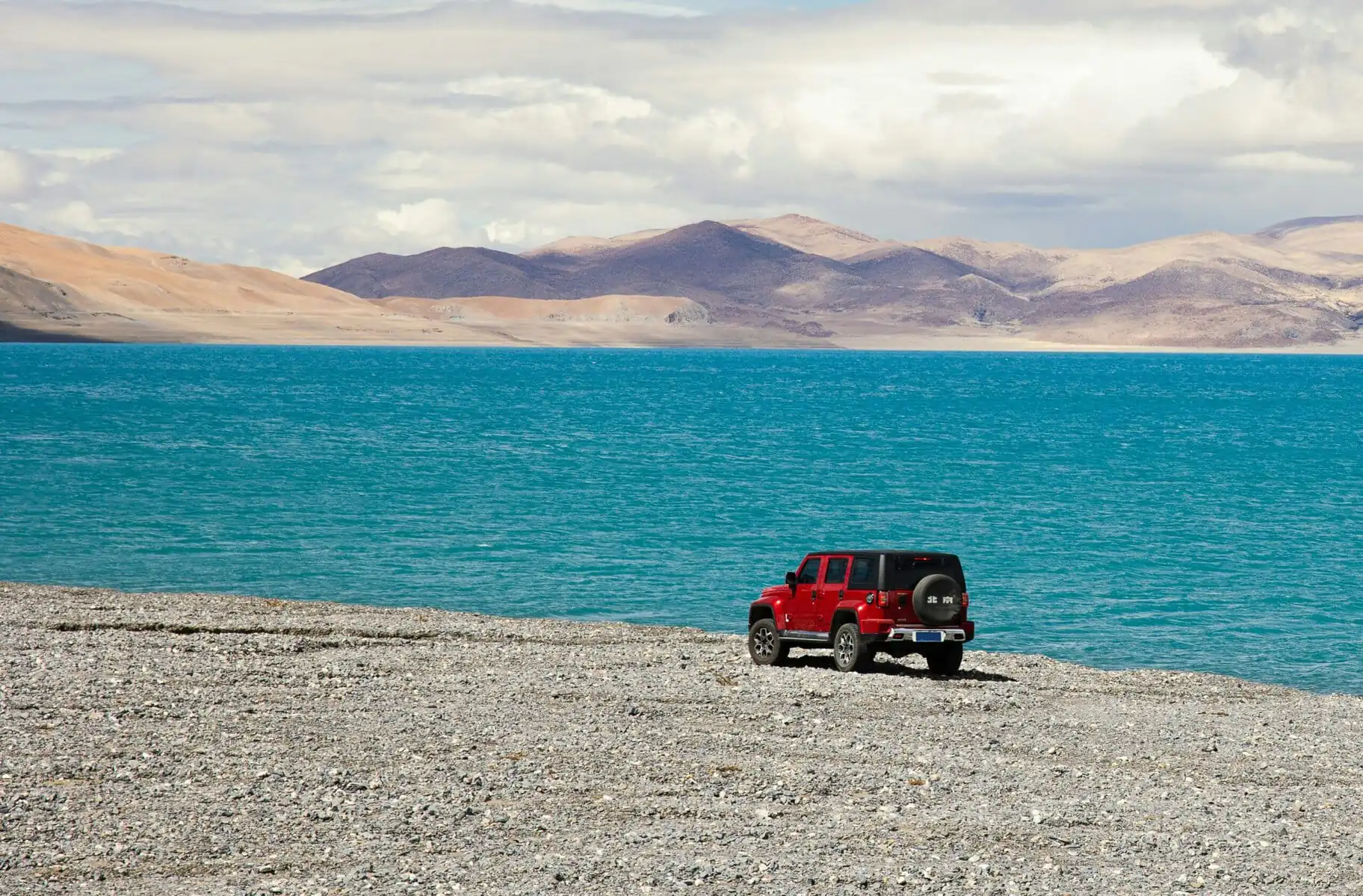 Tibet, china, china tour, Red Jeep parked on a rocky shore beside a bright blue lake with arid mountains in the distance under a cloudy sky