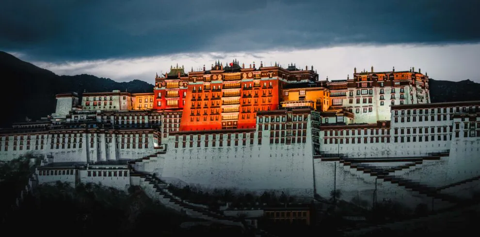 Potala Palace Tibet, china, china tour, Potala Palace perched on terraced hills with warm orange buildings under a dramatic, dark storm sky at dusk.