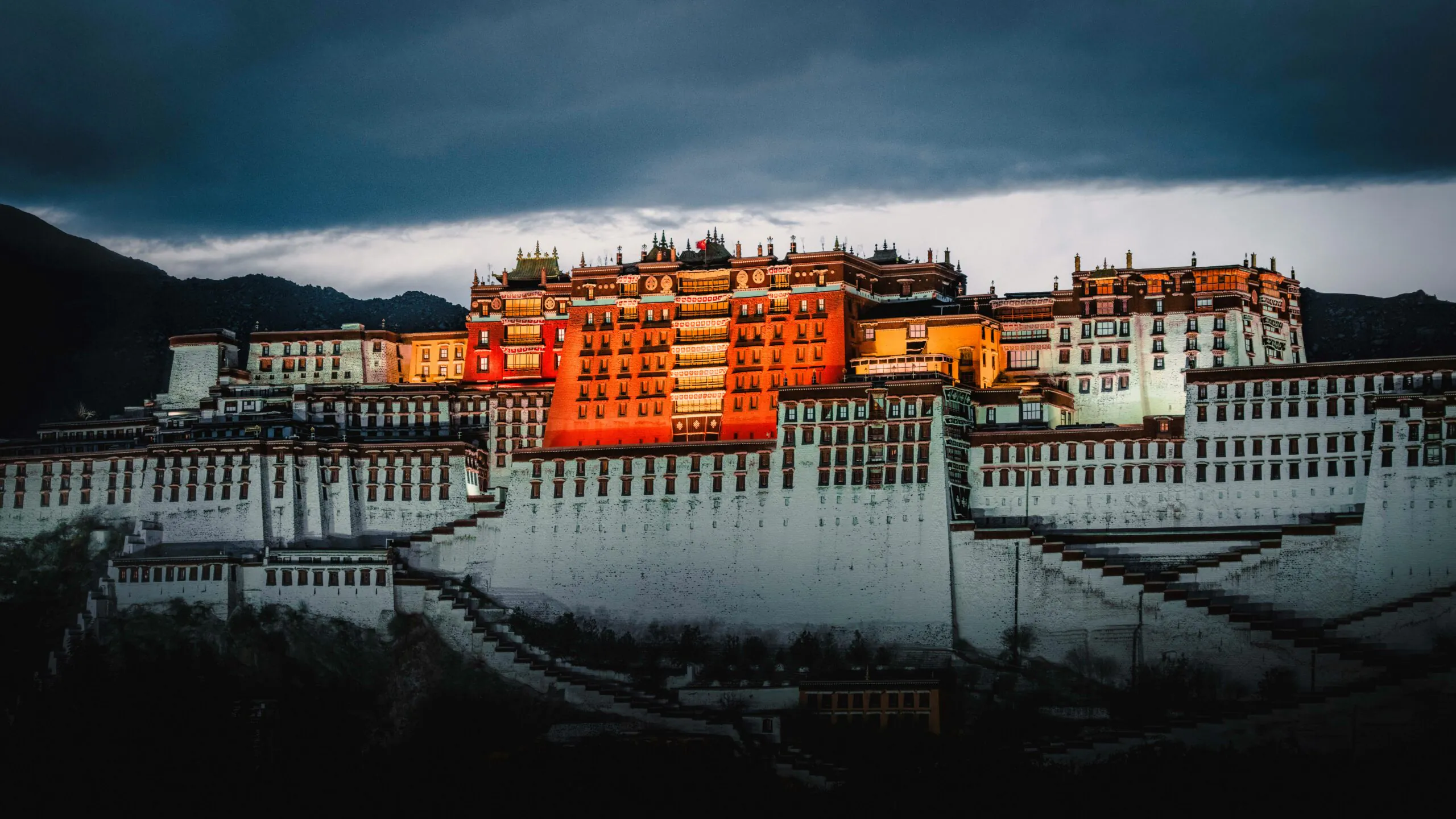 Potala Palace Tibet, china, china tour, Potala Palace perched on terraced hills with warm orange buildings under a dramatic, dark storm sky at dusk.