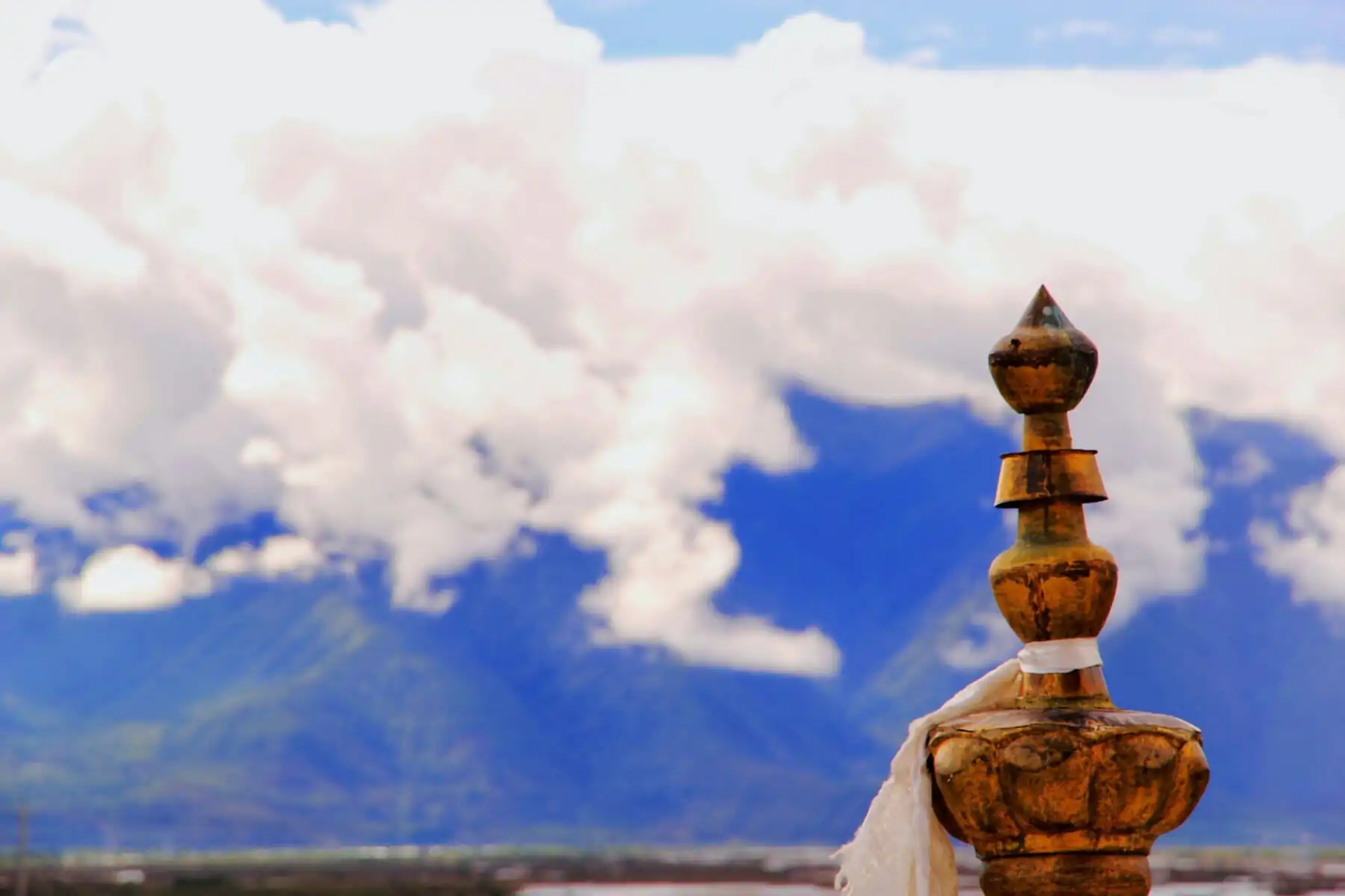 Tibet, china, china tour, Close-up of a weathered wooden finial with a white cloth tied around, set against a blue sky and distant mountains.